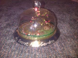 A magical miniature fairy garden depicting a romantic scene where a male fairy floating on a leaf boat on a mystical pond serenades the female fairy that sits playfully in a tree filled with magical fruit surrounded by a garden of wonder. The whole scene plays of in a 22cm glass domed cheese plate
